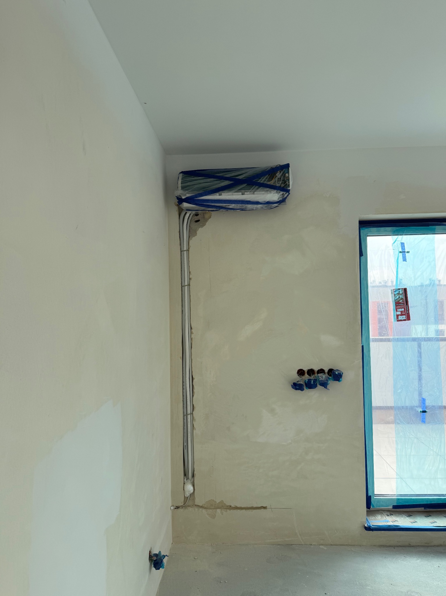 Montaż klimatyzacji w surowym wnętrzu: jednostka wewnętrzna zabezpieczona folią, widoczne rury instalacyjne i przygotowane otwory na gniazdka, okno zakryte folią ochronną.