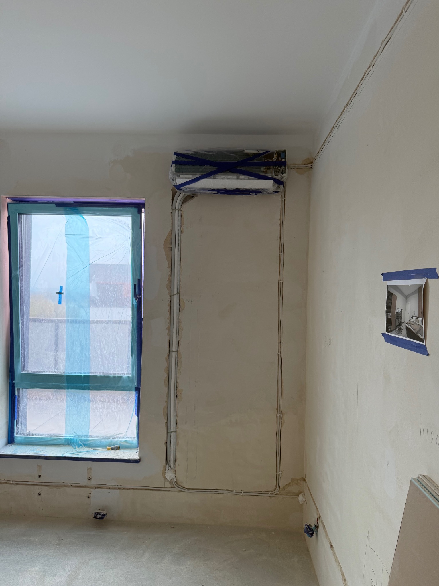 Montaż klimatyzacji w surowym wnętrzu: jednostka wewnętrzna zabezpieczona folią, widoczne przewody instalacyjne na ścianie. Okno zakryte folią ochronną.