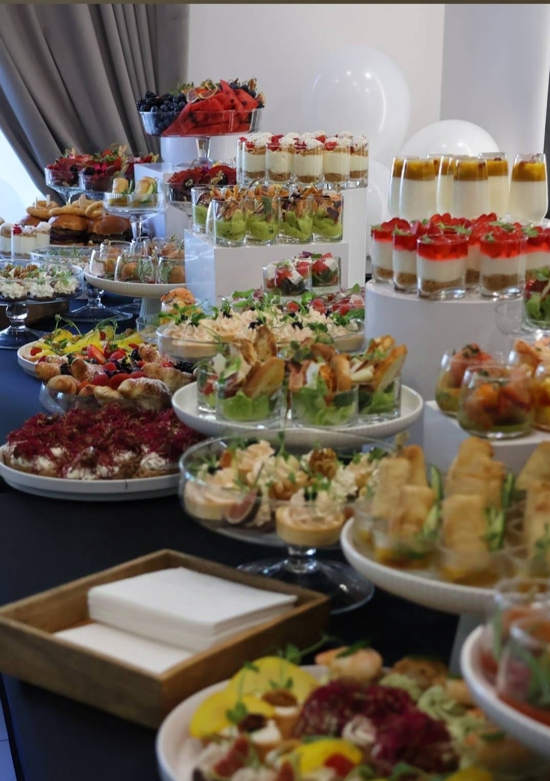 Bogato zastawiony stół z przekąskami i deserami w szklanych naczyniach i na paterach, widoczne owoce, warzywa, ciasta i sałatki; realizacja cateringu w Łodzi, w tle białe balony.