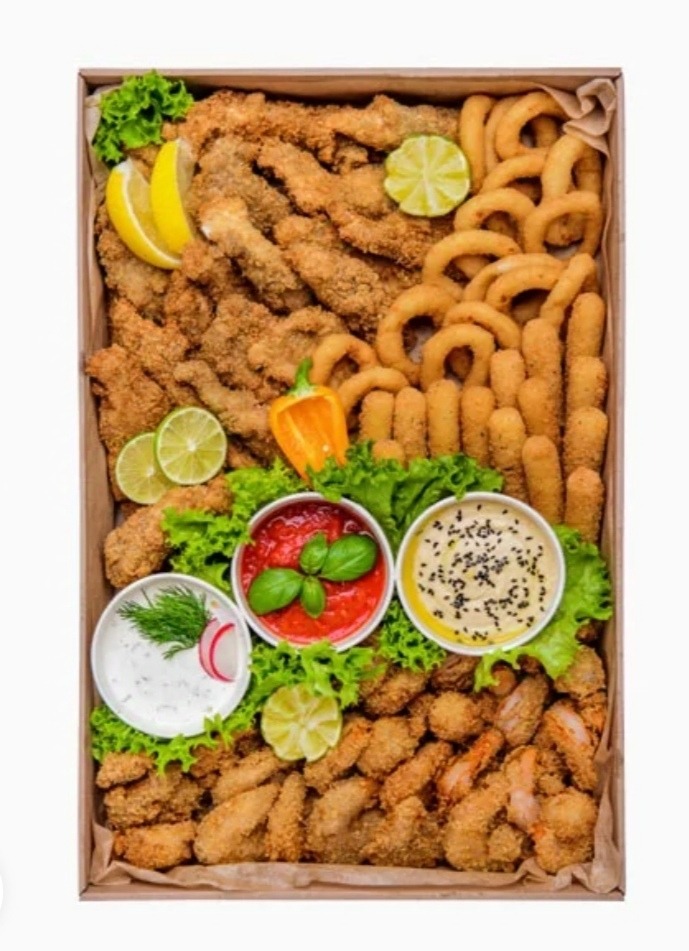 Różnorodne przekąski smażone w głębokim tłuszczu, ułożone w kartonowym pudełku, ozdobione sałatą, plasterkami cytryny i limonki, z trzema miseczkami sosów: czosnkowy, pomidorowy z bazylią i tahini...