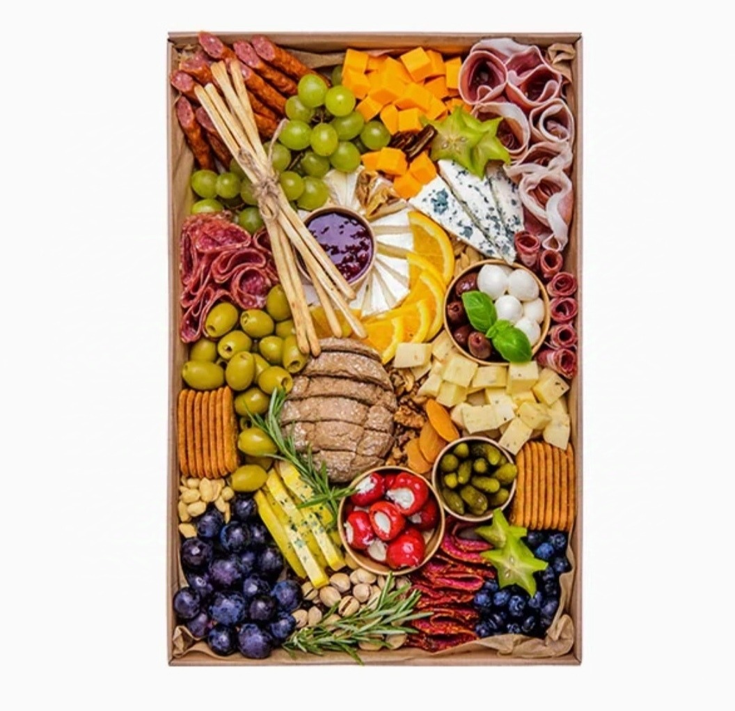 Bogato wypełniona drewniana skrzynka z przekąskami: sery, wędliny, owoce, pieczywo, oliwki, marynowane warzywa, orzechy i dżem, ułożone w dekoracyjny sposób.