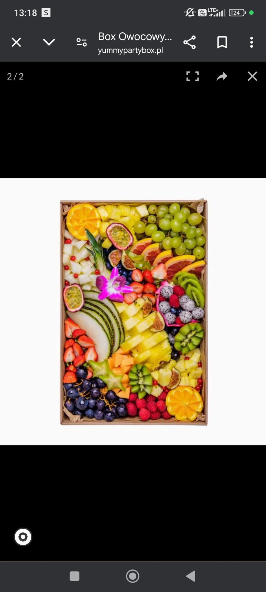 Kompozycja z różnorodnych owoców egzotycznych i sezonowych, ułożonych w drewnianej skrzynce, udekorowana fioletowym kwiatem storczyka.