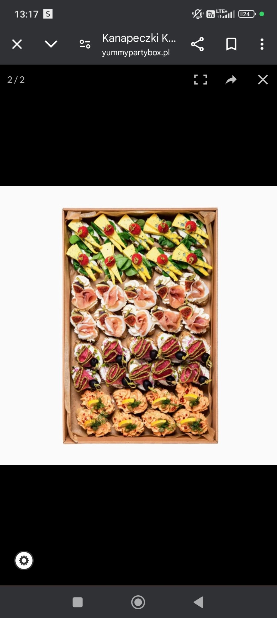 Różnorodne kanapki i przekąski w tekturowym pudełku, widoczne z góry: mini kanapeczki z serem i truskawkami, figami i szynką, mięsem i oliwkami oraz pastą z rybną posypaną koperkiem i cytryną.