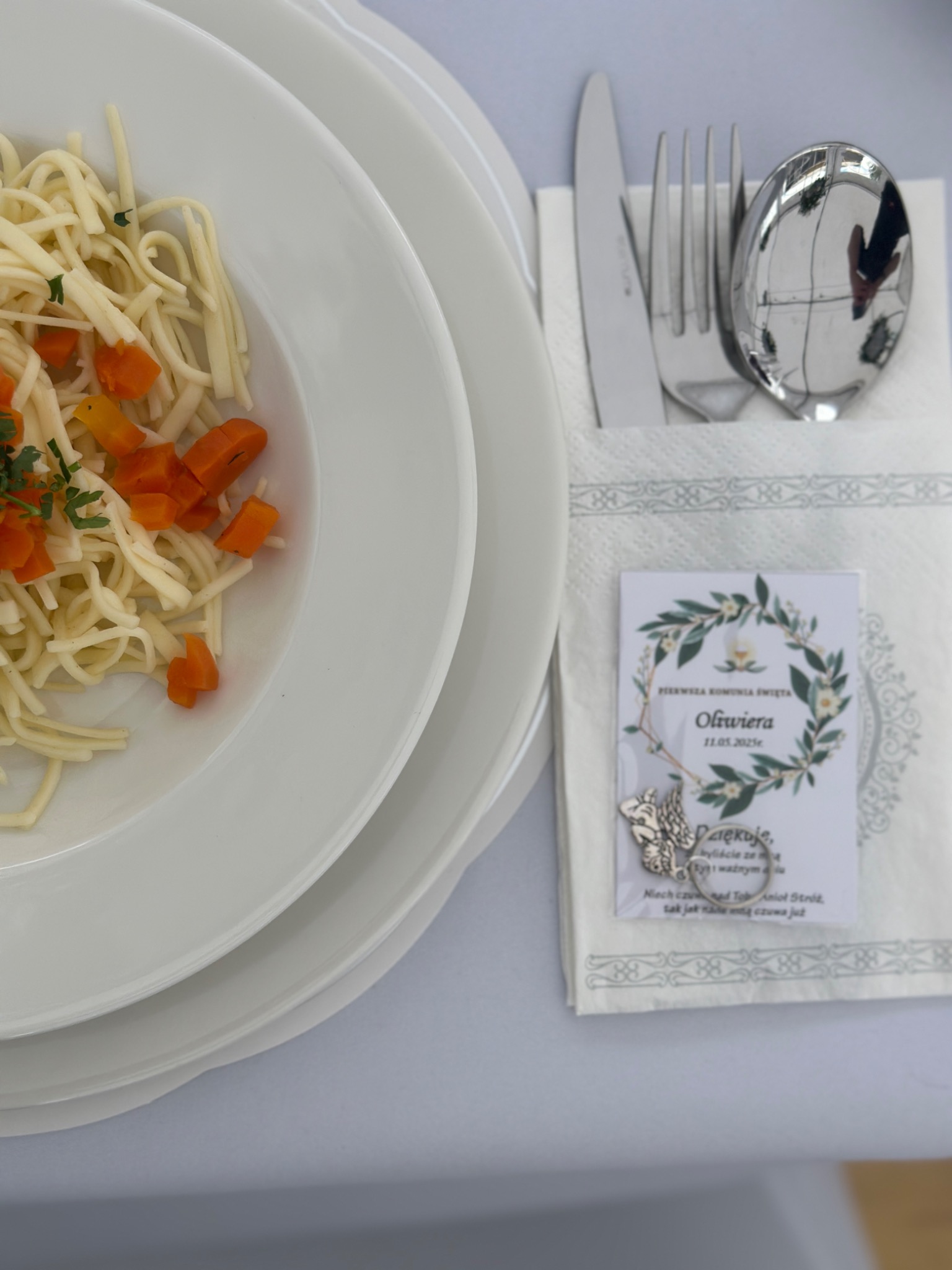 Elegancko nakryty stół: spaghetti z marchewką na białym talerzu, sztućce w serwetce, pamiątka Pierwszej Komunii Świętej, subtelne detale. Uroczysty obiad.