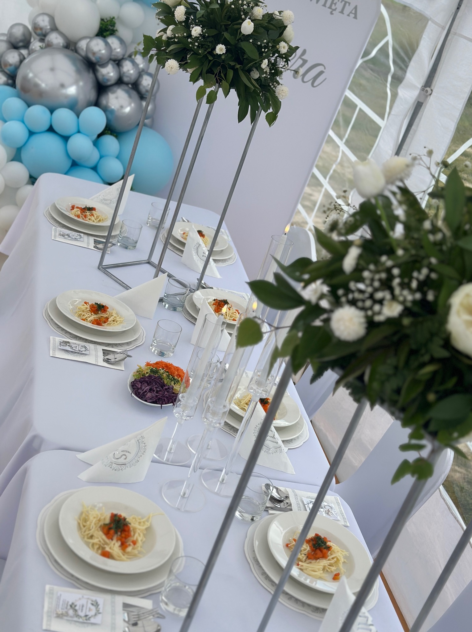 Uroczysty stół z białym obrusem, dekoracjami kwiatowymi, balonami i elegancką zastawą. Na talerzach spaghetti z sosem pomidorowym. Dekoracja sali na przyjęcie.