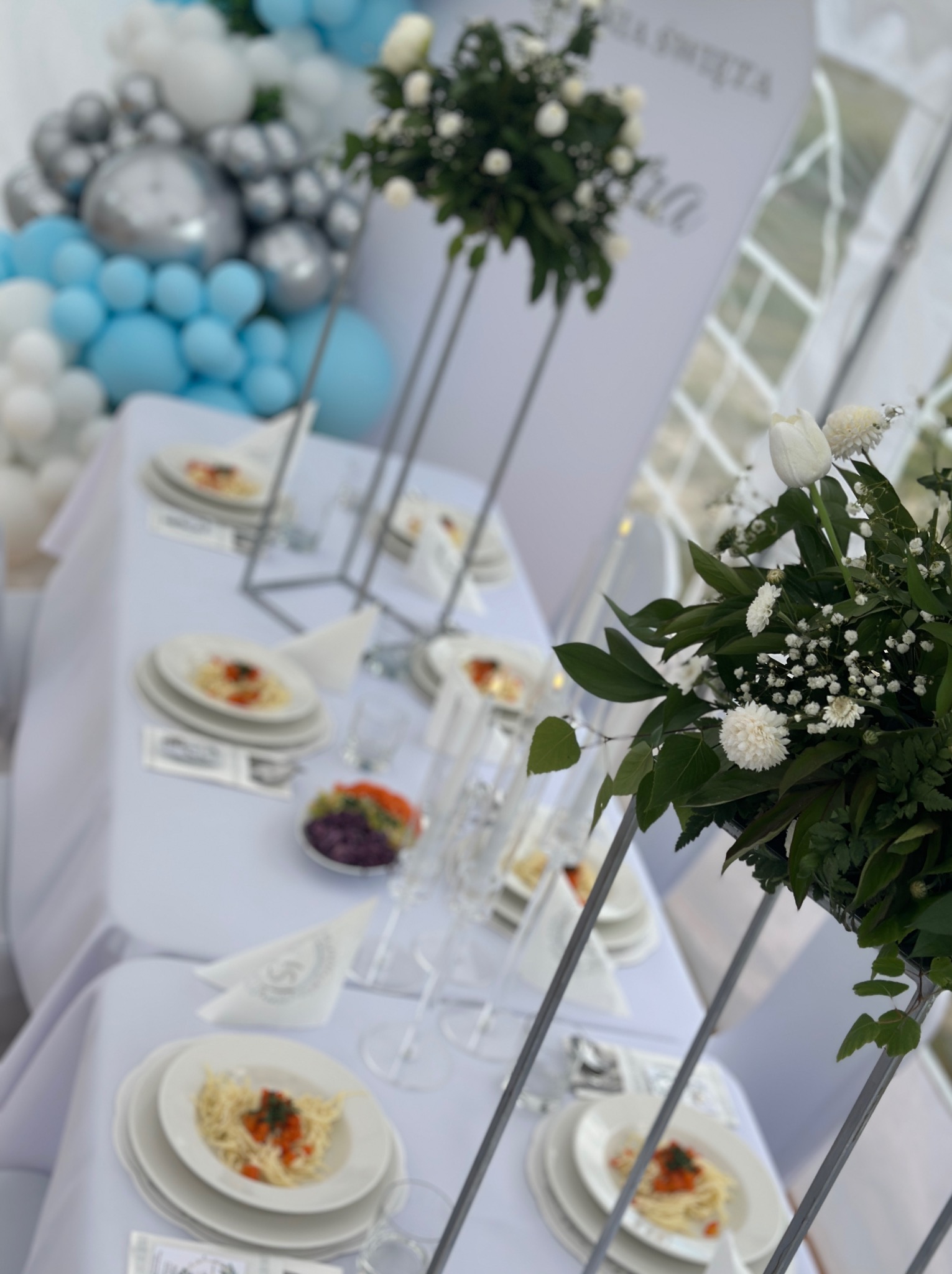 Elegancko nakryty stół z białym obrusem, kwiatami i balonami w odcieniach bieli i błękitu, sugerujący przyjęcie okolicznościowe.