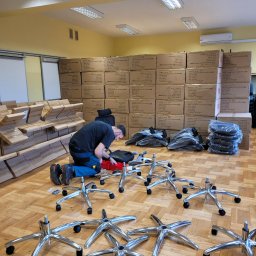Sąsiedzi - Mężczyzna na kolanach składa krzesło biurowe na tle sterty kartonów z krzesłami, widoczne elementy krzesła: podstawy z kółkami, siedziska w folii, narzędzia w czerwonym organizerze.
