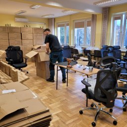 Sąsiedzi - Pracownik w trakcie rozpakowywania biurowych krzeseł z kartonów w przestronnym pomieszczeniu biurowym, z widocznymi narzędziami na stole i stertą złożonych kartonów w tle.