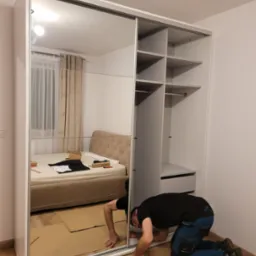 Pracownik w ciemnej koszulce i roboczych spodniach na kolanach, instaluje białą szafę przesuwną z lustrem w sypialni, zabezpieczając podłogę kartonem.