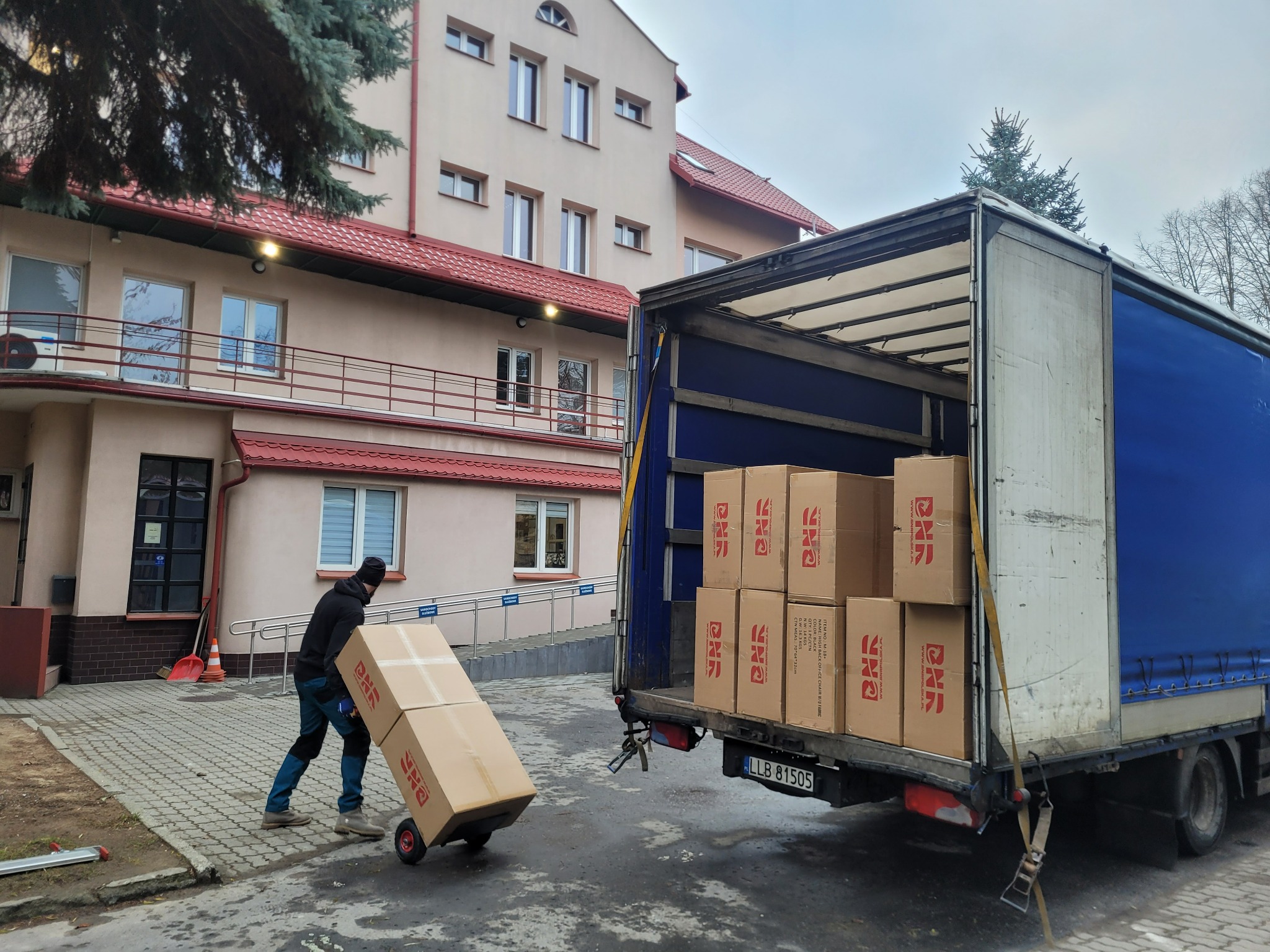 Mężczyzna w czapce i roboczych spodniach transportuje wózeczkiem kartonowe pudło z logo do otwartej ciężarówki z niebieską plandeką, częściowo załadowanej innymi pudłami, na tle budynku z czerwonym...