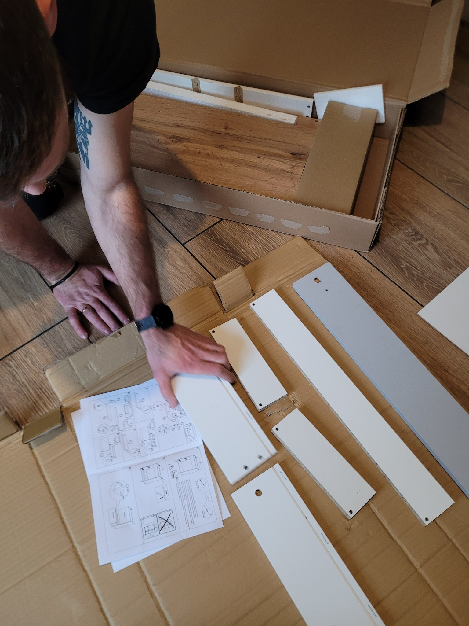 Osoba z tatuażem na ramieniu rozpakowuje elementy mebli z kartonu, rozkładając je na podłodze obok instrukcji montażu.