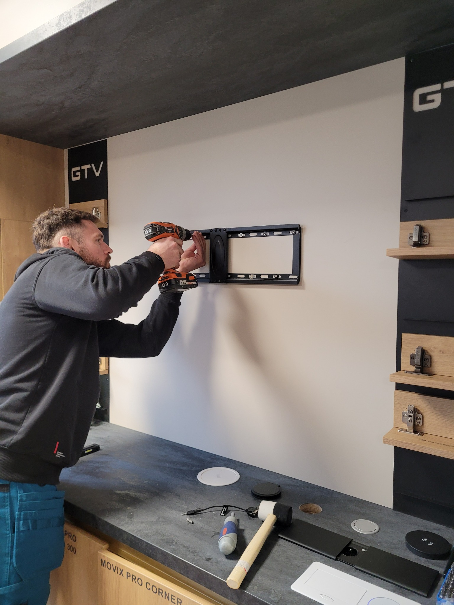 Mężczyzna montuje uchwyt do telewizora na ścianie za pomocą wiertarki AEG, widoczne narzędzia na blacie roboczym, logo GTV w tle.