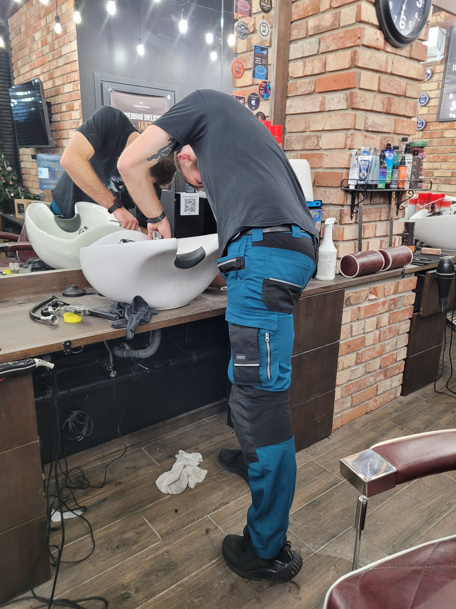 Mężczyzna w spodniach roboczych instaluje umywalkę fryzjerską w salonie, widoczne narzędzia, rury i elementy instalacyjne.