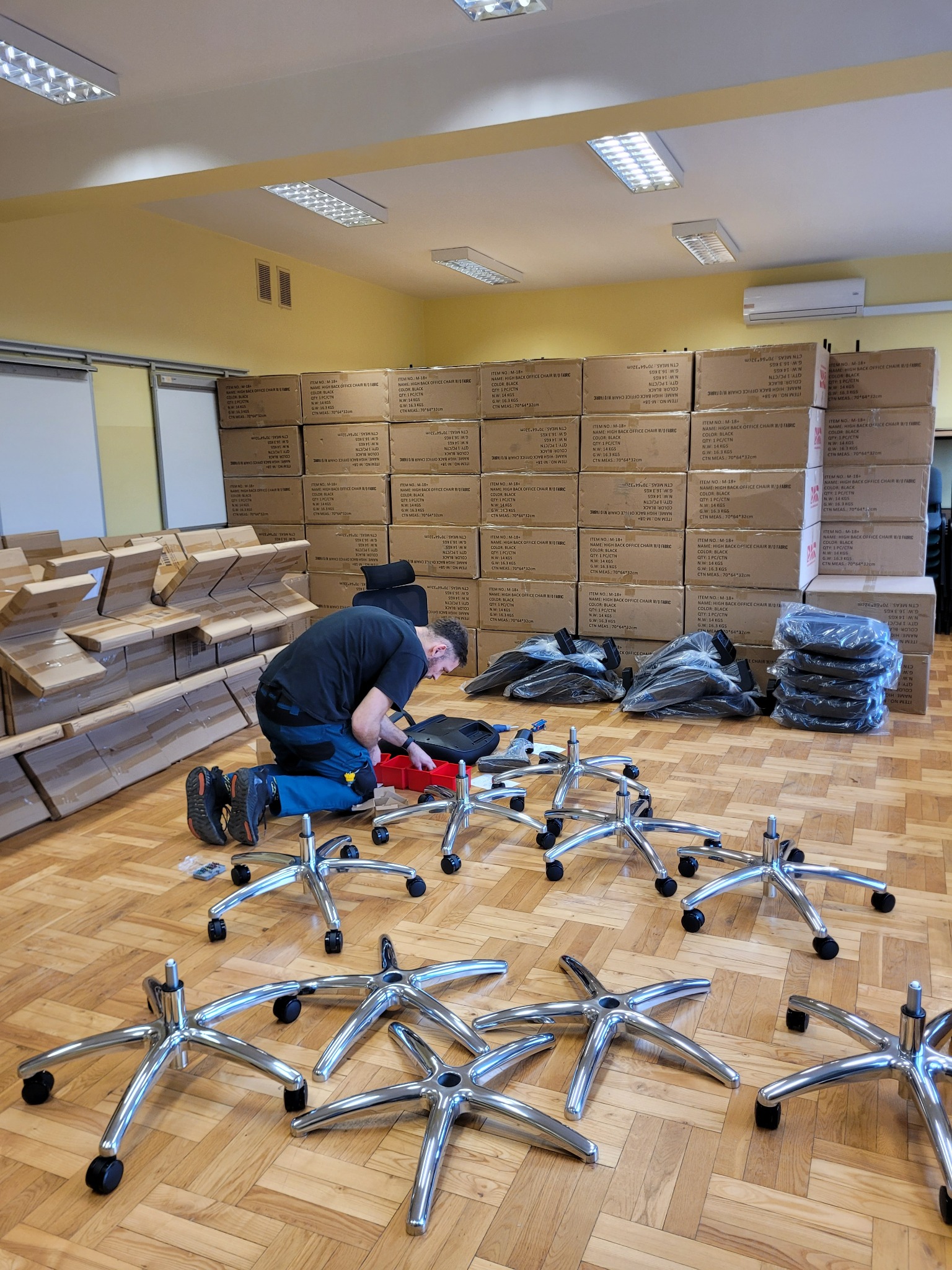 Mężczyzna na kolanach składa krzesło biurowe na tle sterty kartonów z krzesłami, widoczne elementy krzesła: podstawy z kółkami, siedziska w folii, narzędzia w czerwonym organizerze.