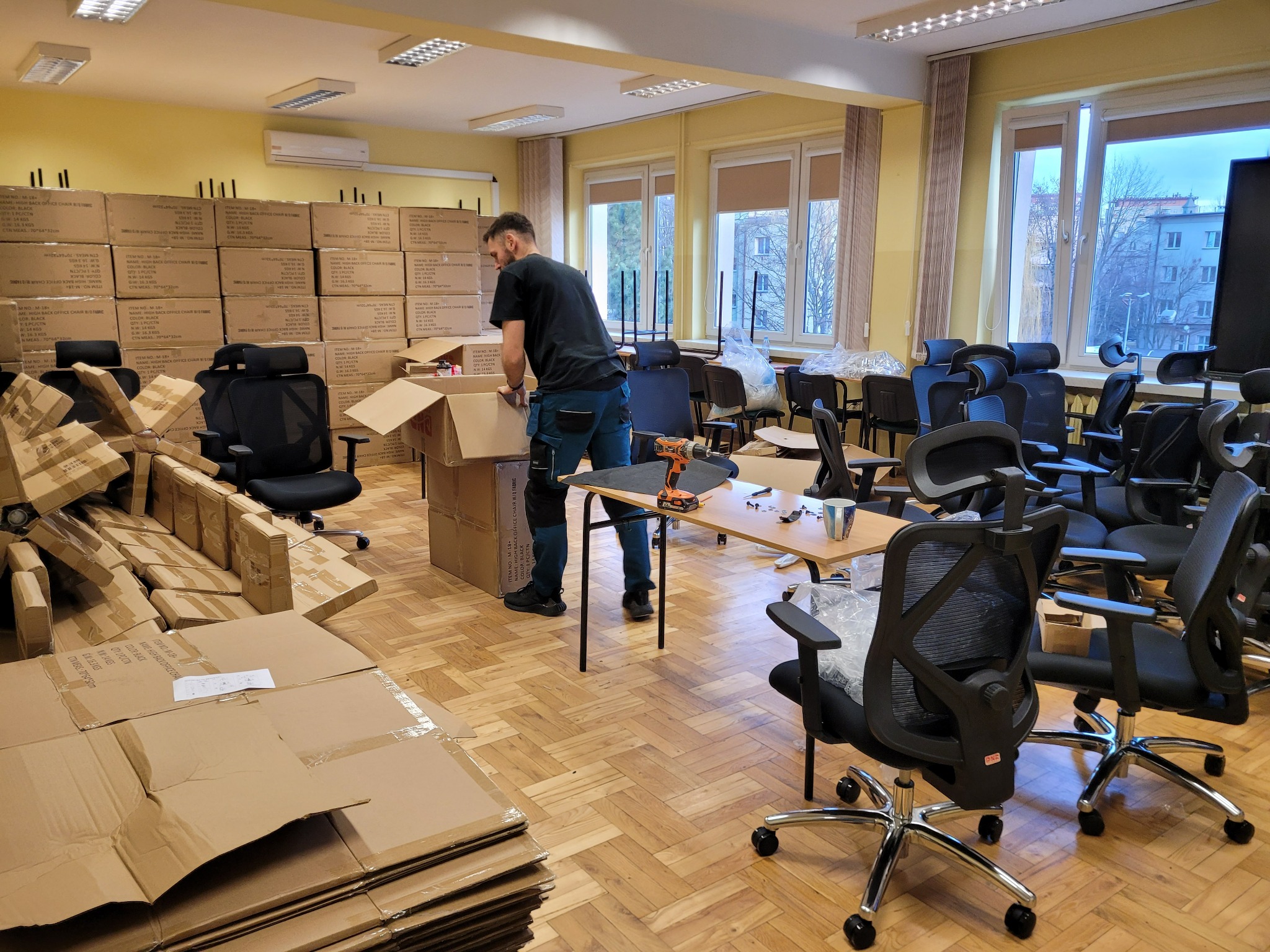 Pracownik w trakcie rozpakowywania biurowych krzeseł z kartonów w przestronnym pomieszczeniu biurowym, z widocznymi narzędziami na stole i stertą złożonych kartonów w tle.