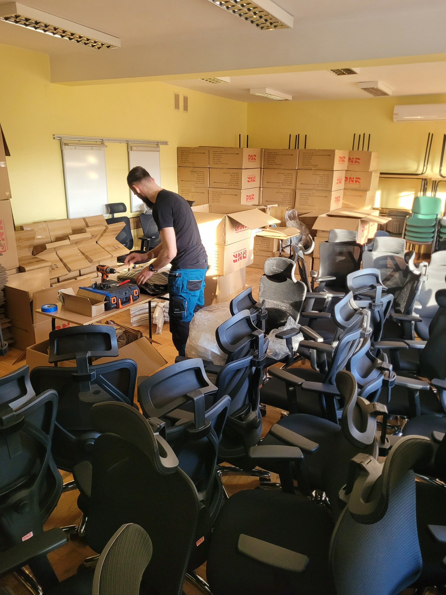 Mężczyzna w niebieskich spodniach roboczych montuje czarne krzesła biurowe w pomieszczeniu z żółtymi ścianami, narzędzia i pudełka w tle.