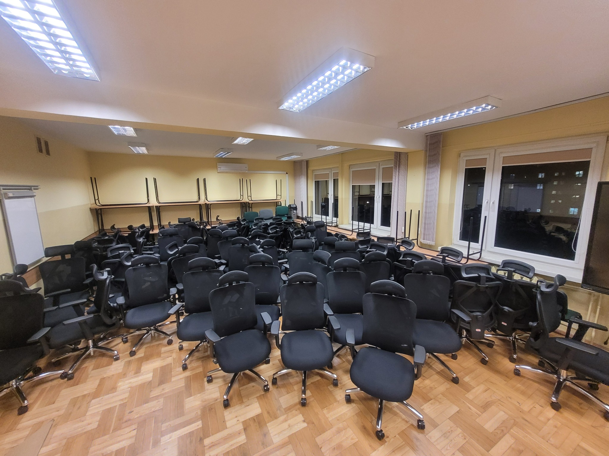 Sala konferencyjna z dużą ilością czarnych krzeseł biurowych na kółkach, ustawionych na parkiecie w jodełkę, z biurkami i krzesłami ustawionymi przy ścianach pod oknami, oświetlona przez kasetonowe...