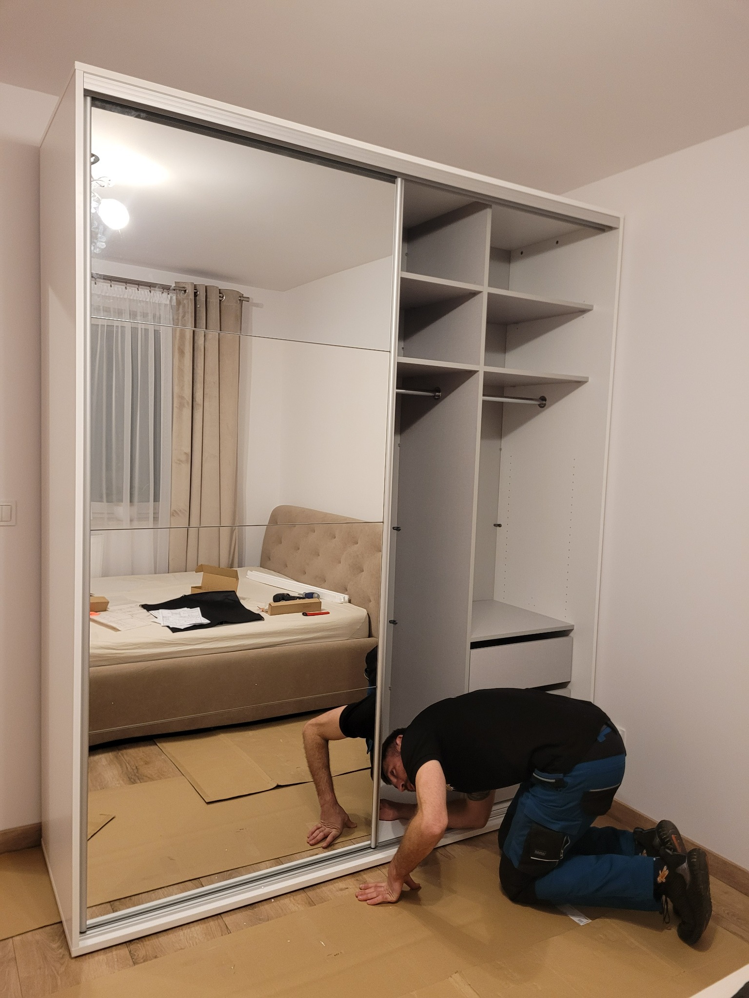 Pracownik w ciemnej koszulce i roboczych spodniach na kolanach, instaluje białą szafę przesuwną z lustrem w sypialni, zabezpieczając podłogę kartonem.