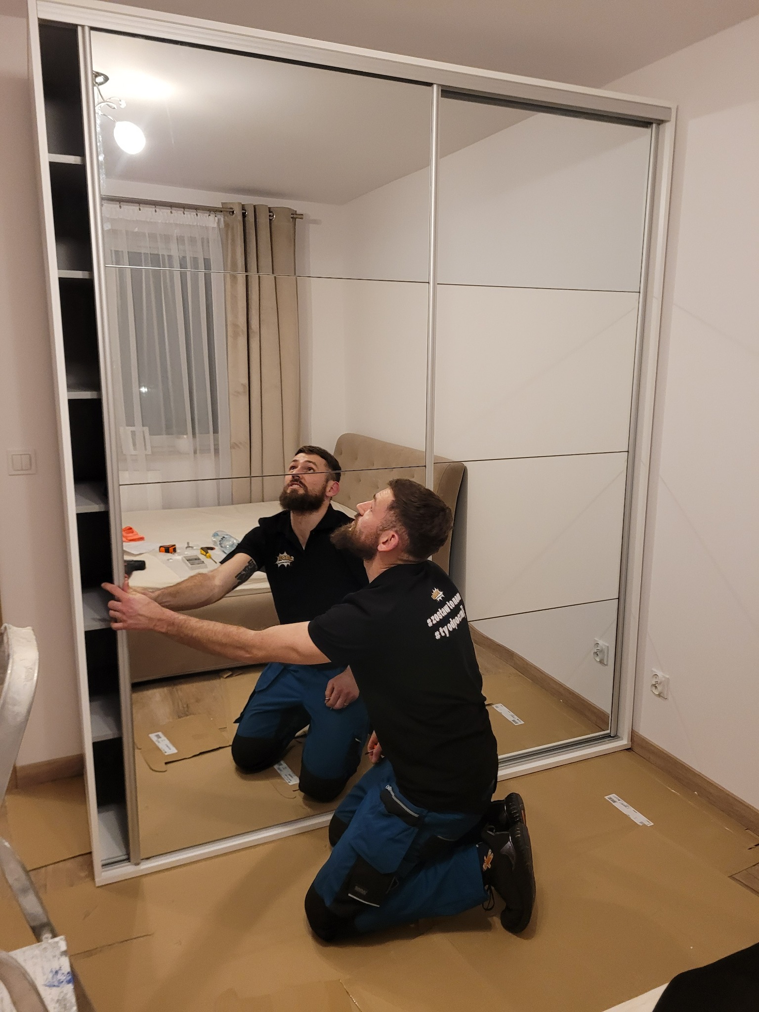 Dwóch monterów na kolanach instaluje lustrzane drzwi przesuwne do szafy wnękowej, zabezpieczając podłogę kartonem. Widoczne narzędzia i częściowo złożona konstrukcja szafy.