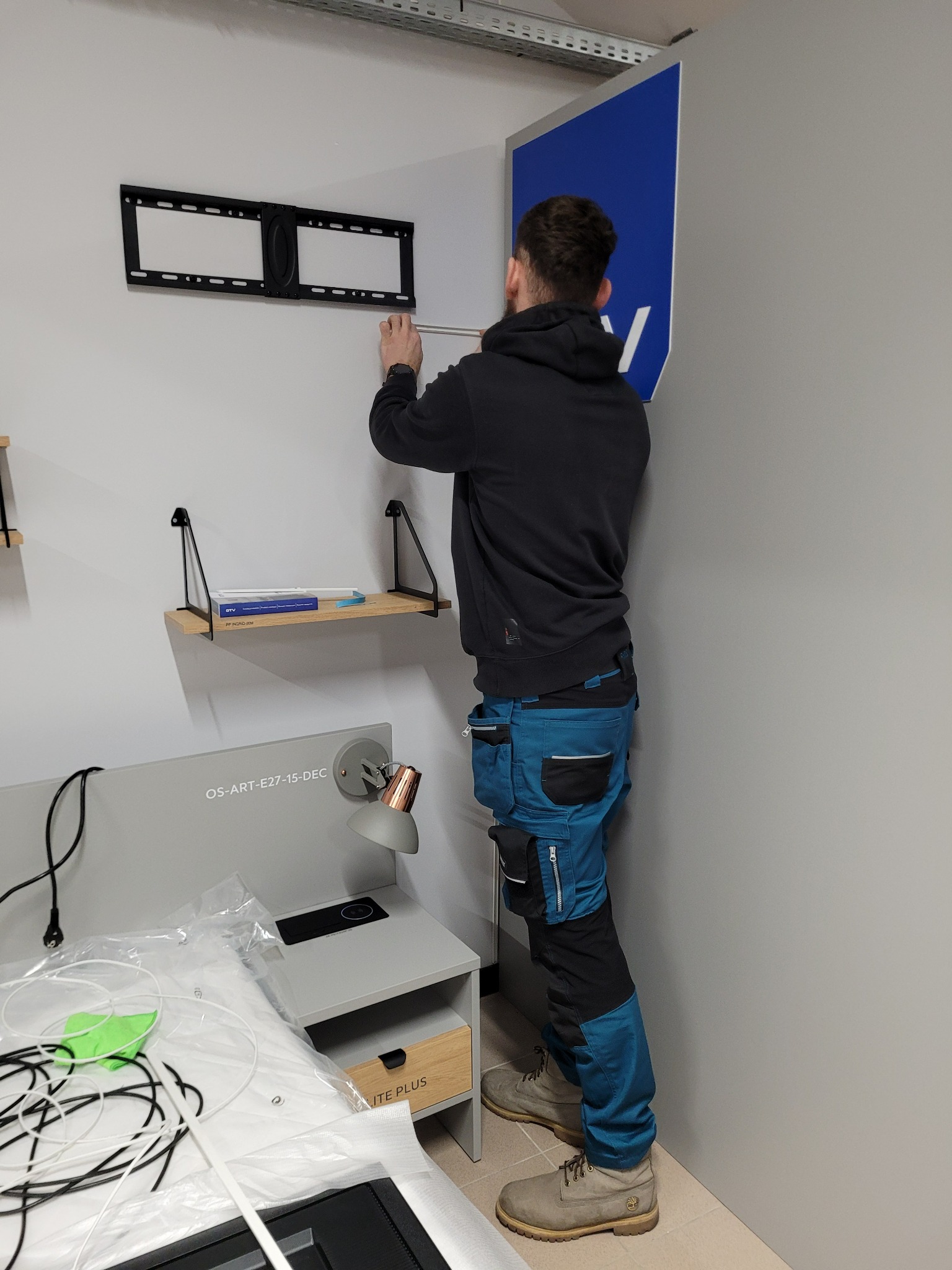 Mężczyzna w roboczych spodniach montuje uchwyt ścienny do telewizora w nowoczesnym wnętrzu, widoczny stolik nocny z lampką i półki.