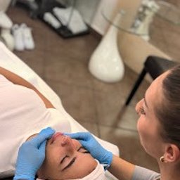 Art Clinik Kraków - Chirurgia Plastyczna - Medycyna Estetyczna - Kosmetologia - Kobieta leżąca na leżance podczas zabiegu na twarz wykonywanego przez kosmetyczkę w niebieskich rękawiczkach; widoczne oświetlenie punktowe i szklany stolik w tle.