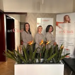 Trzy kobiety w uniformach stojące w holu placówki medycznej, za nimi plakat reklamowy z logo 'art Clinik' i kwiaty w białej donicy.