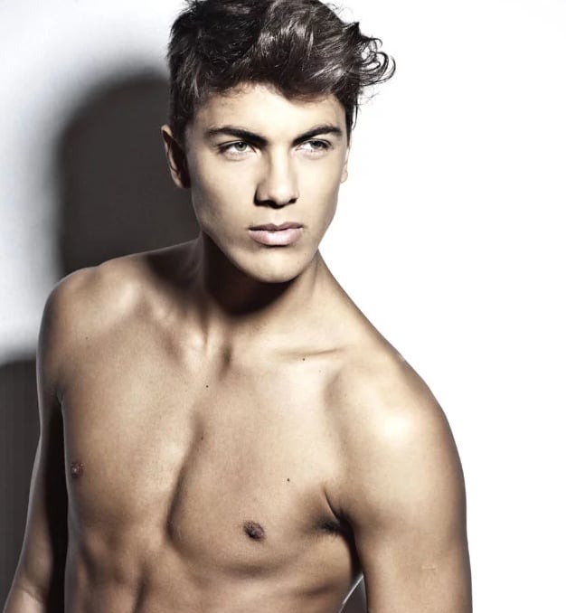 Portret młodego mężczyzny z krótkimi, ciemnymi włosami i zielonymi oczami, na białym tle, z widoczną klatką piersiową i ramionami.