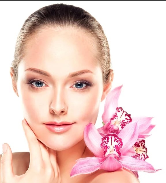 Portret kobiety z delikatnym makijażem, dotykającej policzka, z różowymi orchideami przy ramieniu na białym tle.