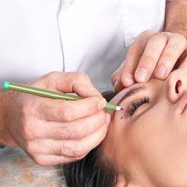 Osoba leżąca podczas zabiegu, zaznaczanie konturu oka zielonym flamastrem, widoczne sztuczne rzęsy.