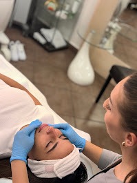 Kobieta leżąca na leżance podczas zabiegu na twarz wykonywanego przez kosmetyczkę w niebieskich rękawiczkach; widoczne oświetlenie punktowe i szklany stolik w tle.