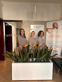 Trzy kobiety w uniformach stojące w holu placówki medycznej, za nimi plakat reklamowy z logo 'art Clinik' i kwiaty w białej donicy.