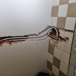 Ja siam - Przygotowanie do instalacji armatury w łazience: widoczne rury z czerwonymi złączkami wystające ze ściany, częściowo usunięte płytki i odsłonięta surowa ściana.