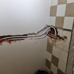 Przygotowanie do instalacji armatury w łazience: widoczne rury z czerwonymi złączkami wystające ze ściany, częściowo usunięte płytki i odsłonięta surowa ściana.