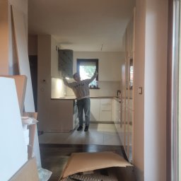 Ja siam - Wnętrze mieszkania w trakcie prac wykończeniowych, widoczne kartony i materiały budowlane, w tle osoba w kuchni z podniesionymi rękami, zamontowany okap, minimalistyczne białe szafki.