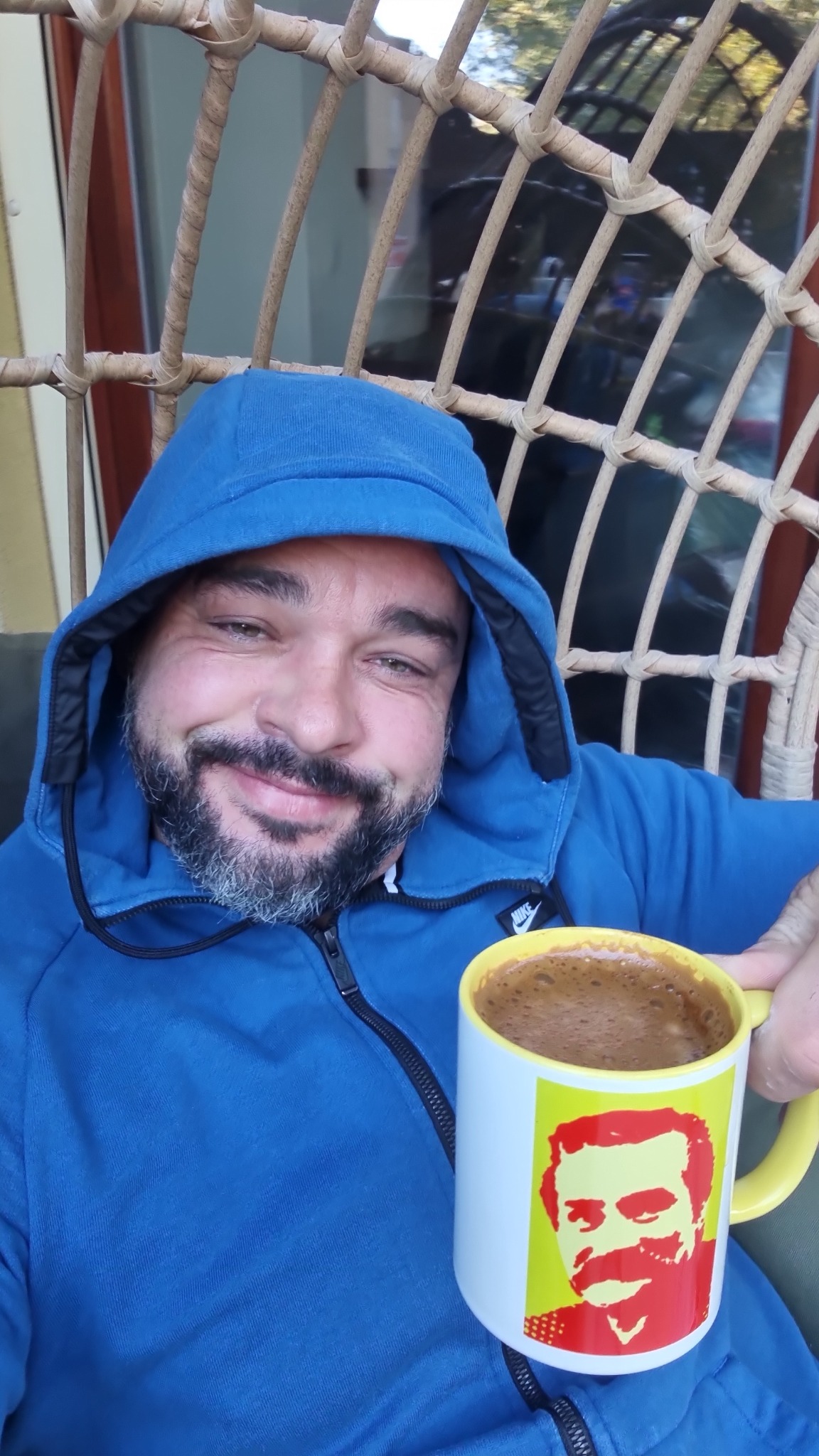 Uśmiechnięty mężczyzna z siwą brodą w niebieskiej bluzie z kapturem, siedzący w wiklinowym fotelu, trzyma kubek z kawą z portretem mężczyzny.