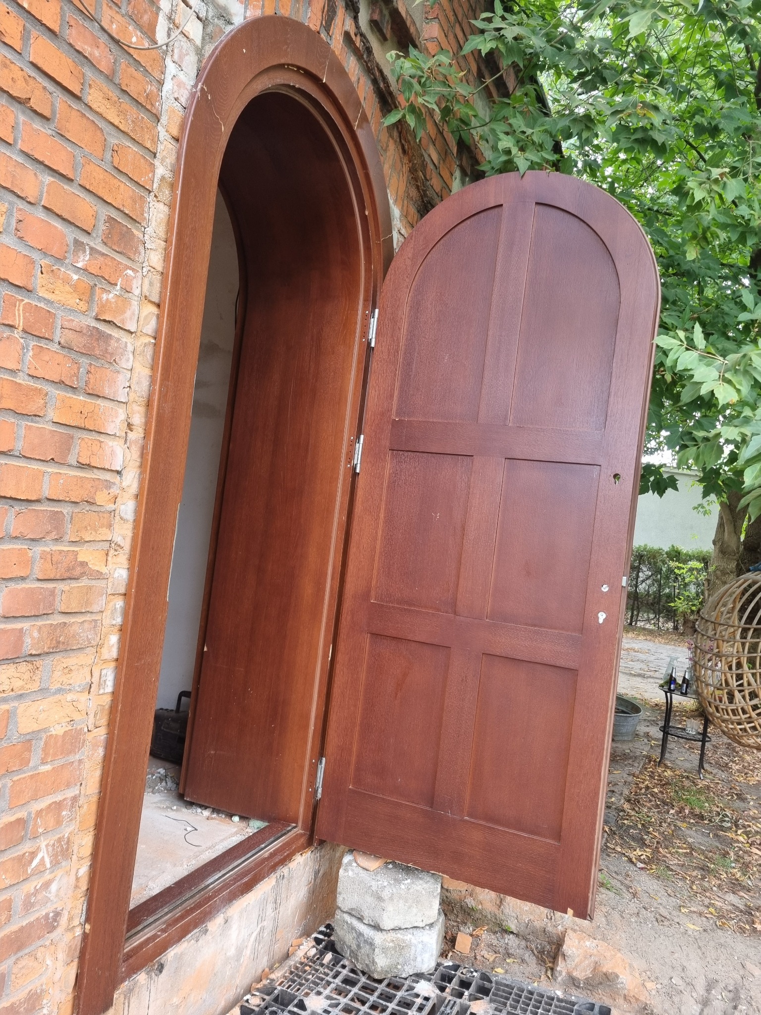 Otwarta, brązowa, drewniana brama z zaokrąglonym łukiem w murze z czerwonej cegły, widoczne zawiasy i fragmentarycznie podłoże z kratką ściekową.