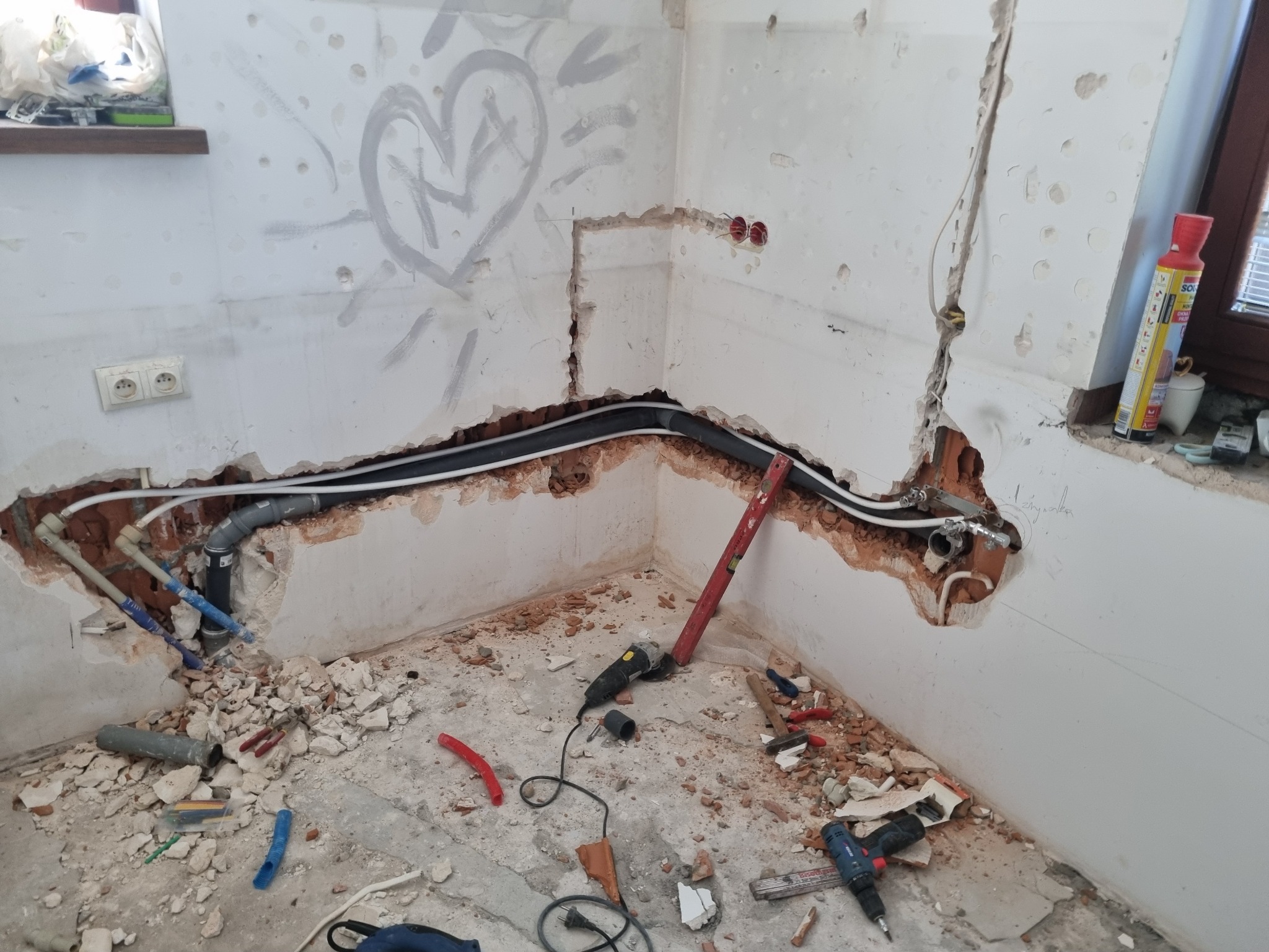 Ściana w trakcie prac instalacyjnych z widocznymi rurami, przewodami elektrycznymi, gruzem i narzędziami, na ścianie widnieje amatorski rysunek serca.