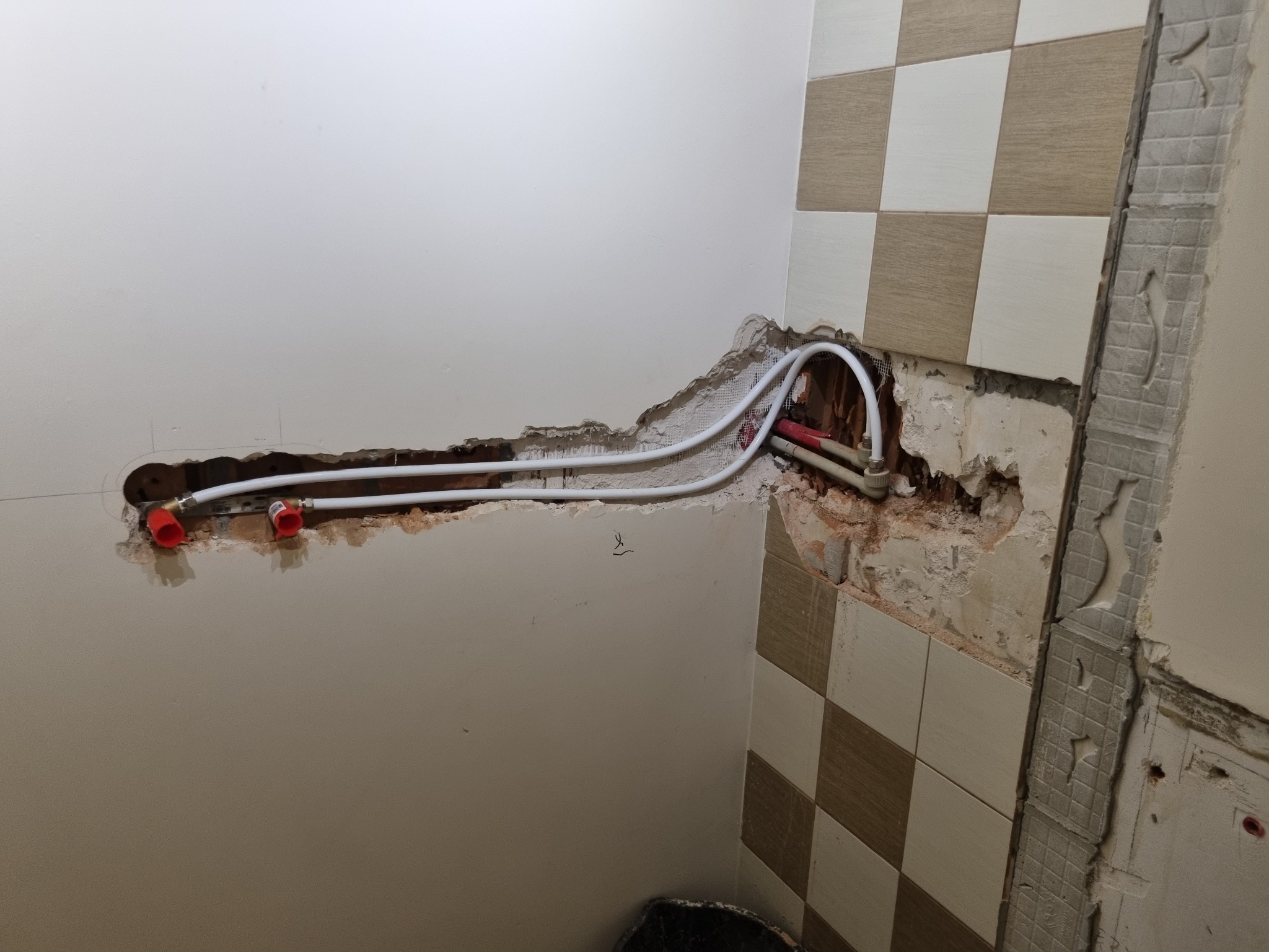 Przygotowanie do instalacji armatury w łazience: widoczne rury z czerwonymi złączkami wystające ze ściany, częściowo usunięte płytki i odsłonięta surowa ściana.