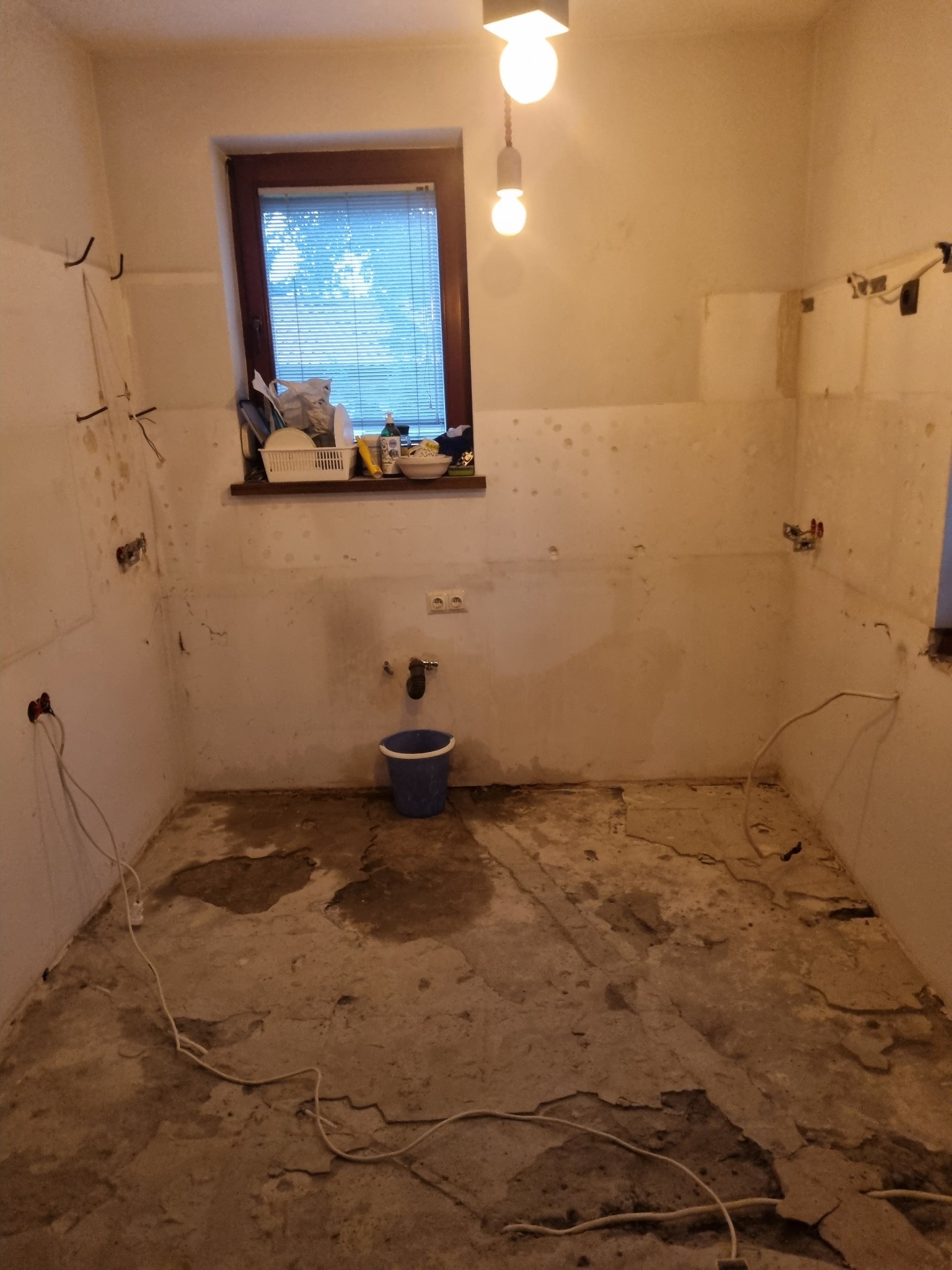 Łazienka w trakcie demontażu starych płytek, widoczne odsłonięte ściany i podłoga, wiszące kable elektryczne, okno z żaluzjami i wiadro na podłodze.