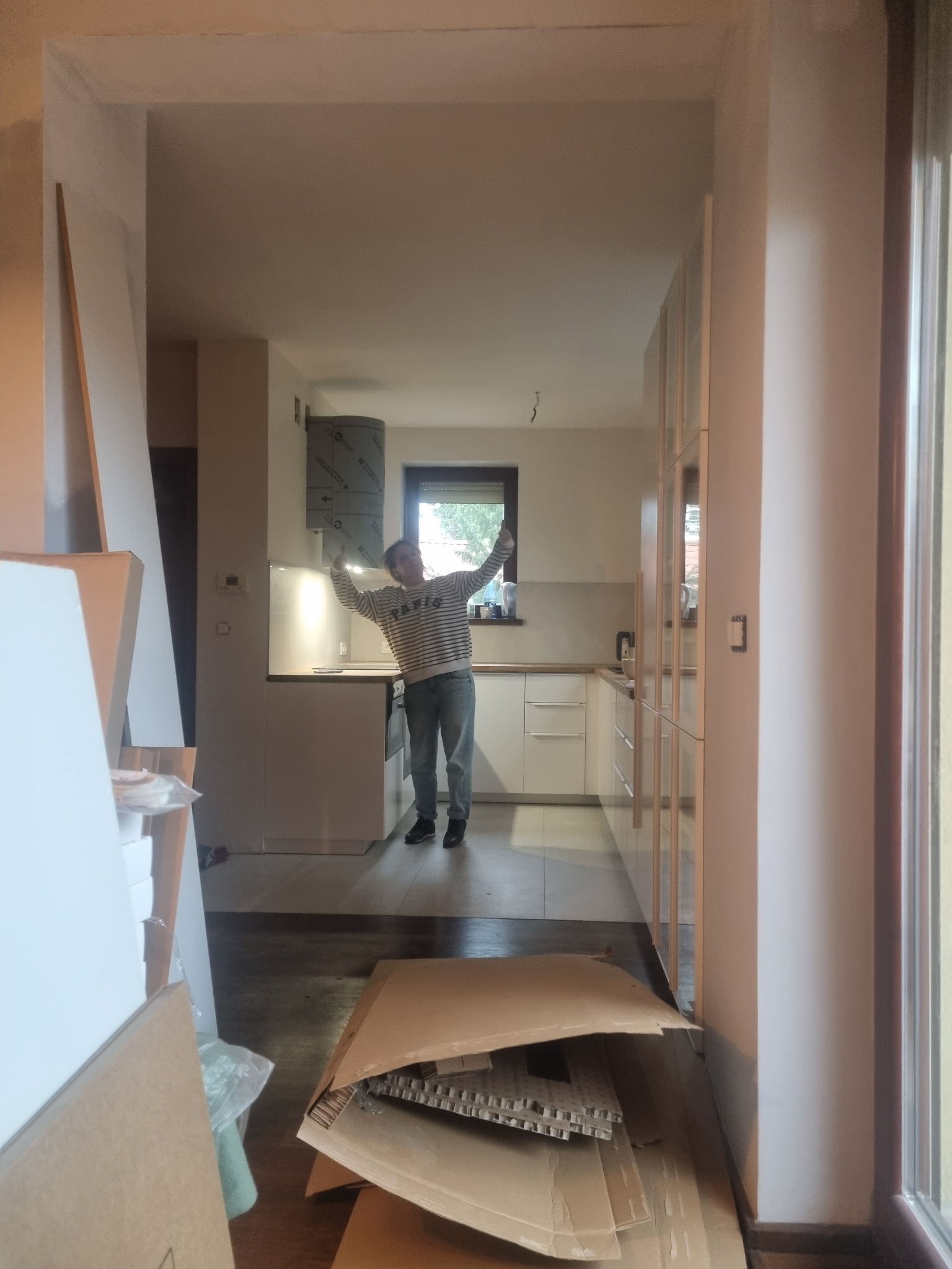 Wnętrze mieszkania w trakcie prac wykończeniowych, widoczne kartony i materiały budowlane, w tle osoba w kuchni z podniesionymi rękami, zamontowany okap, minimalistyczne białe szafki.