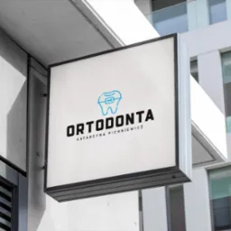 Kwadratowy, podświetlany szyld reklamowy z logo gabinetu ortodontycznego, zamontowany na elewacji nowoczesnego budynku.