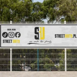 Billboard reklamowy z logotypem Street Unity, adres strony internetowej i ikonami social media, umieszczony na ogrodzeniu z siatki na tle zieleni.