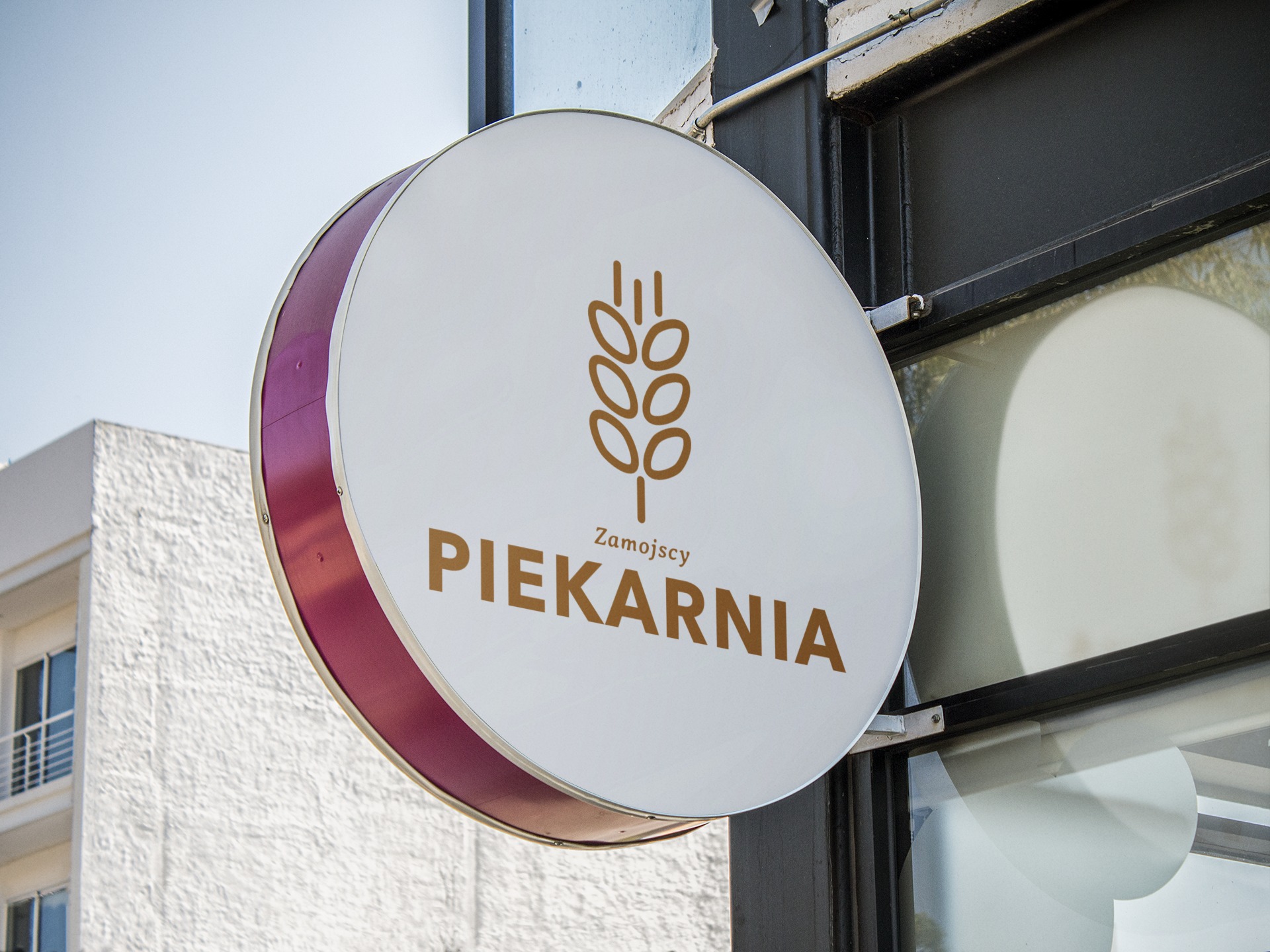 Okrągły szyld piekarni Zamoyscy z grafiką kłosa zboża w kolorze brązowym, umieszczony na zewnątrz budynku z widoczną elewacją w tle.