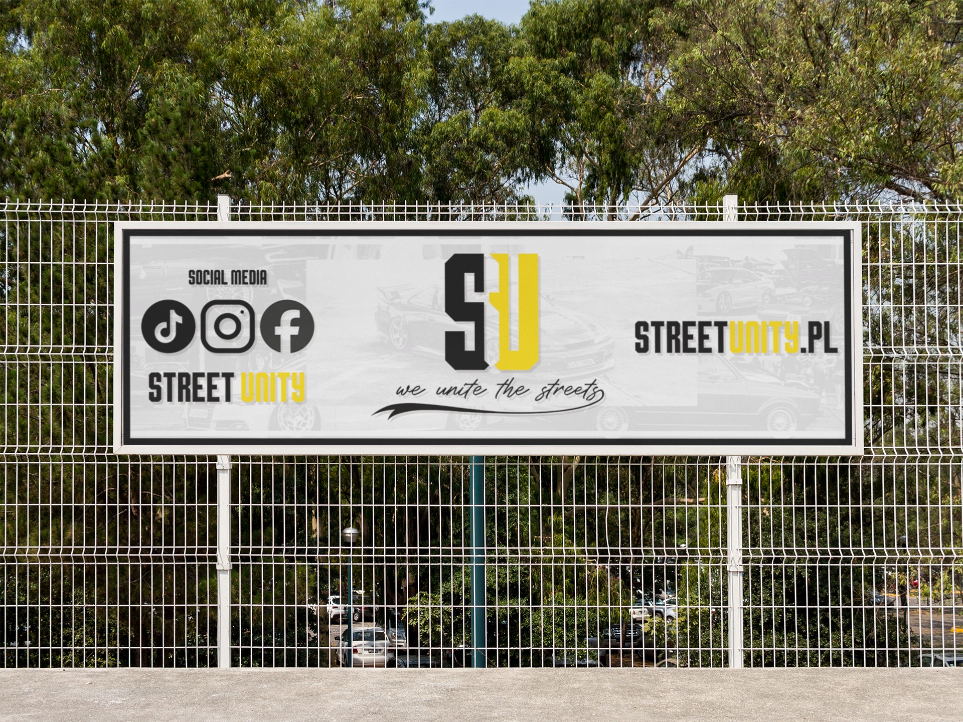 Billboard reklamowy z logotypem Street Unity, adres strony internetowej i ikonami social media, umieszczony na ogrodzeniu z siatki na tle zieleni.