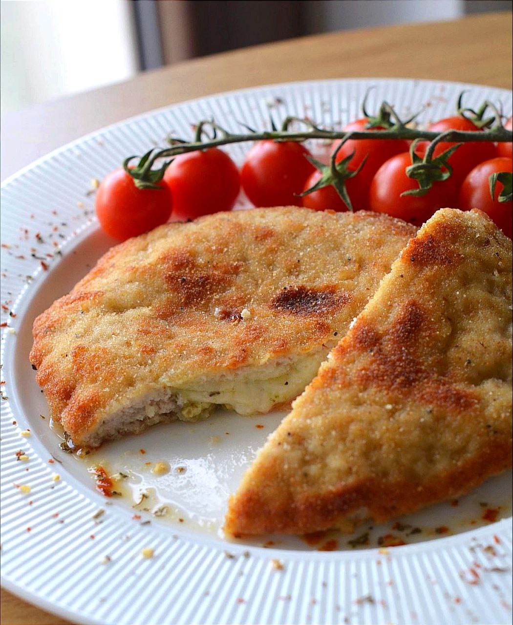 Panierowany sznycel z roztopionym serem w środku, podany z gałązką pomidorków cherry na białym talerzu z dekoracyjnymi żłobieniami.