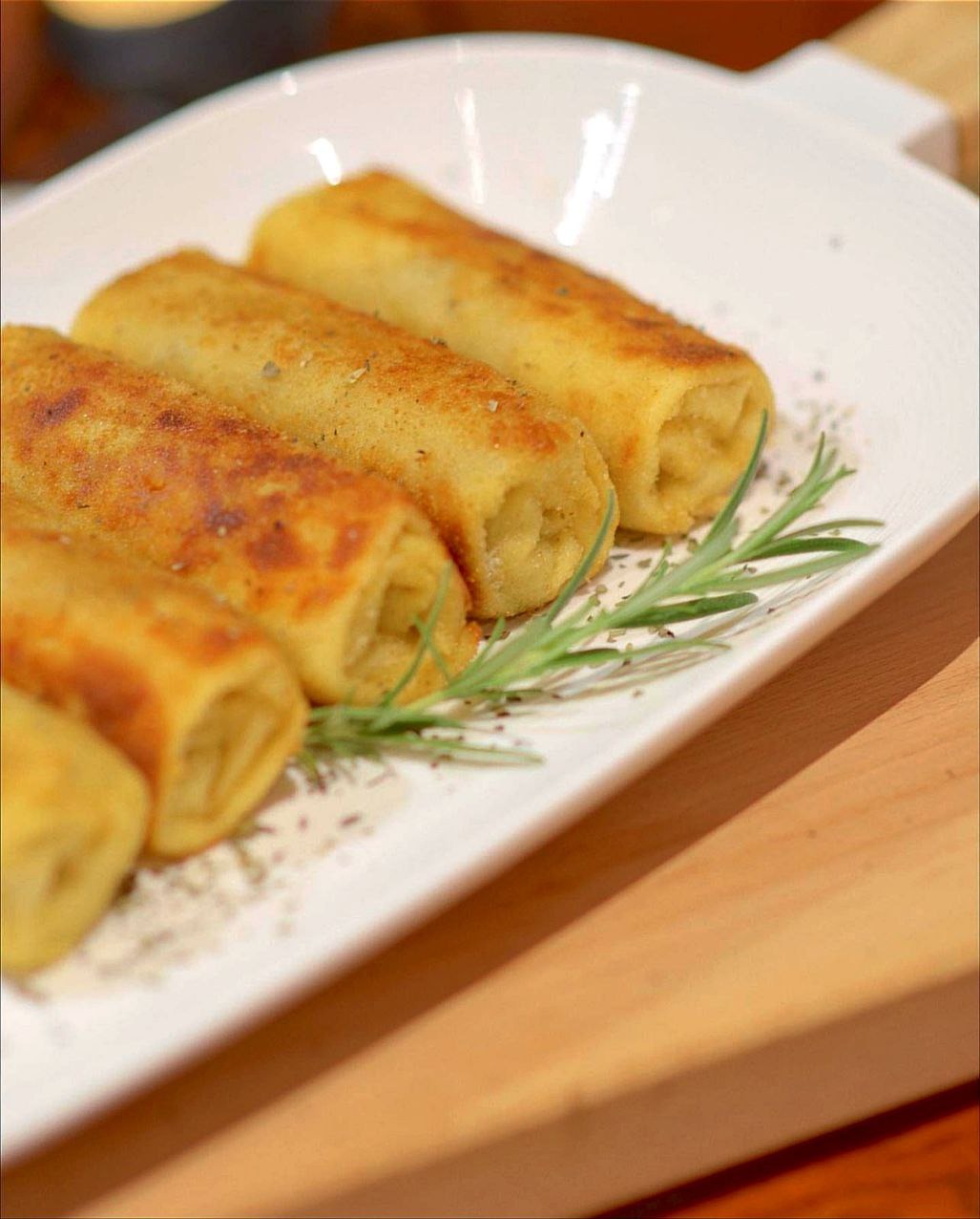 Cztery krokiety ułożone na białym, prostokątnym talerzu, udekorowane gałązką rozmarynu i posypane przyprawami, widoczne na drewnianym blacie.