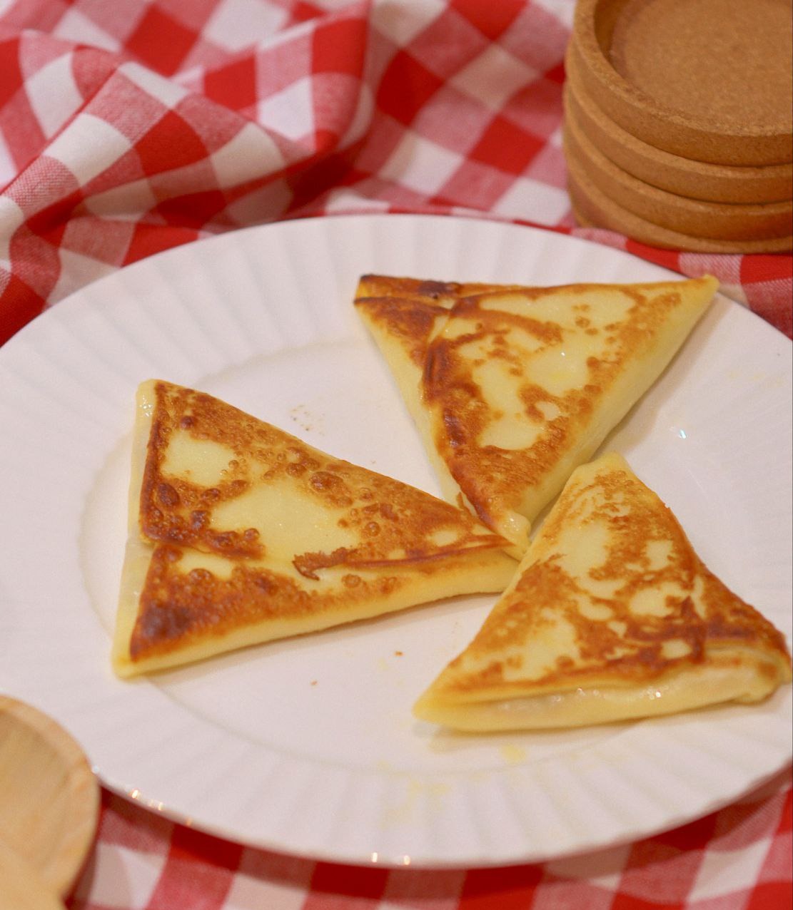Trzy trójkątne naleśniki z serem na białym talerzu, w tle czerwono-biała krata i drewniane podkładki.