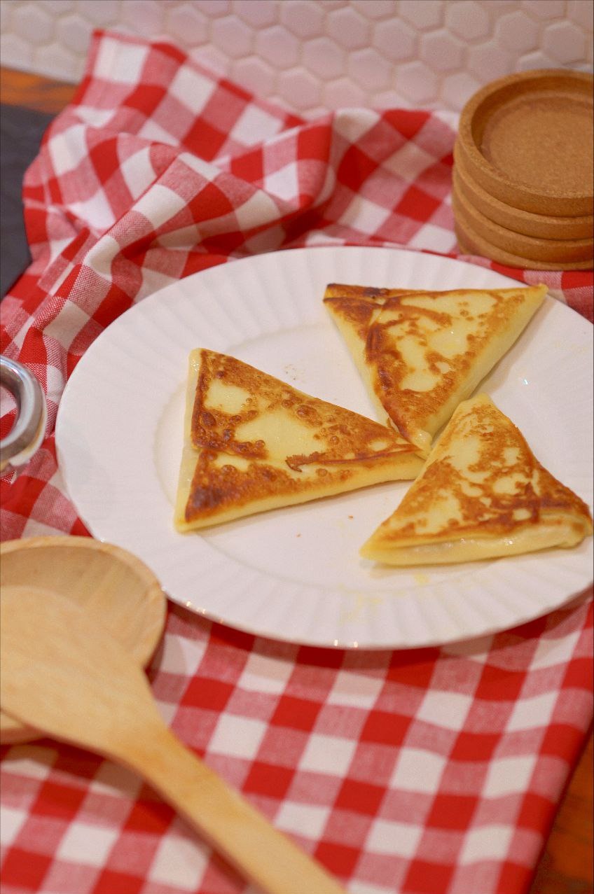 Trzy naleśniki w kształcie trójkątów, ułożone na białym talerzu z karbowanym brzegiem, na tle czerwono-białej kraty i drewnianej łyżki.