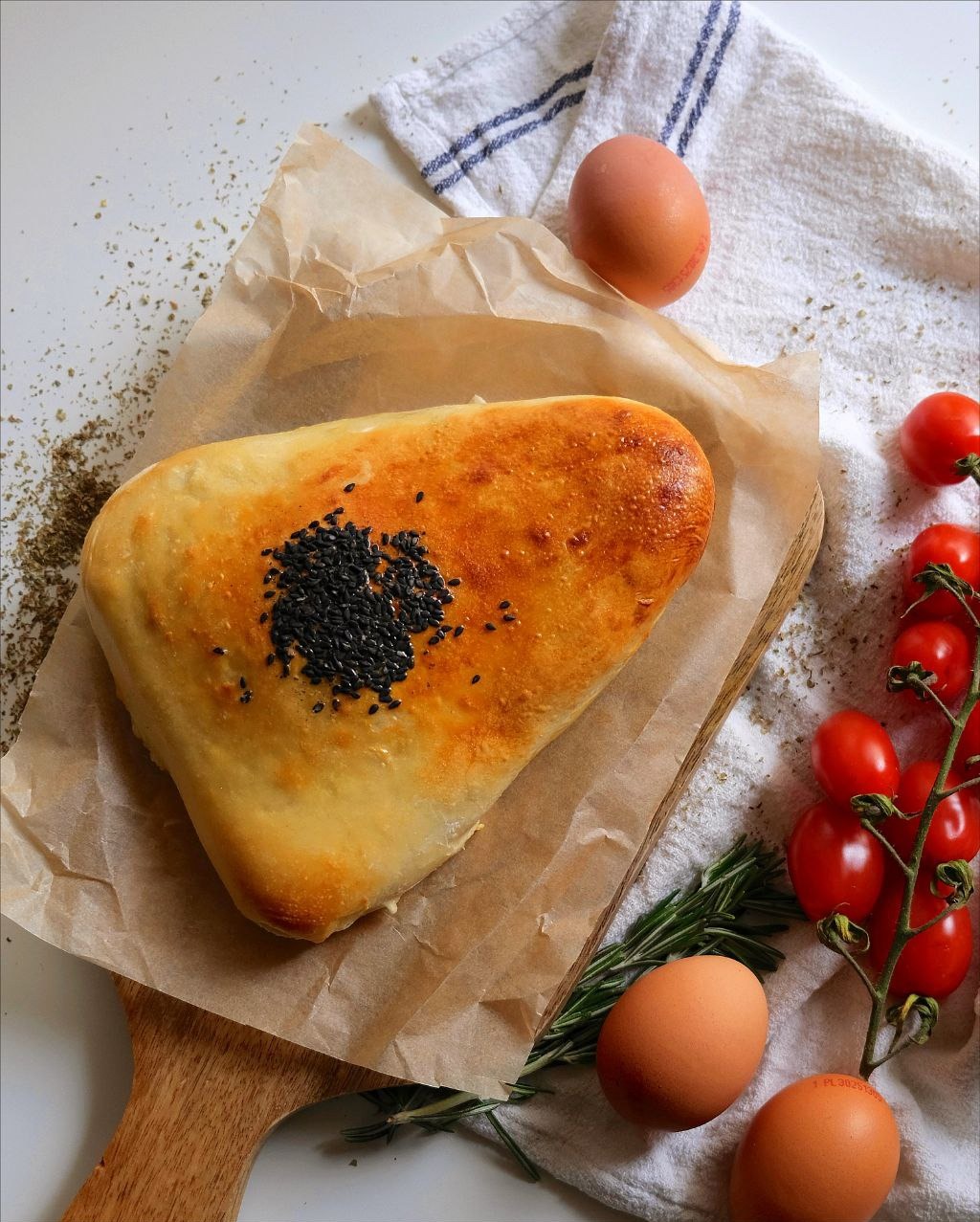 Domowe ciasto z czarnuszką na pergaminie, drewniana deska, gałązka pomidorów koktajlowych, jajka i biała ściereczka z niebieskimi paskami.