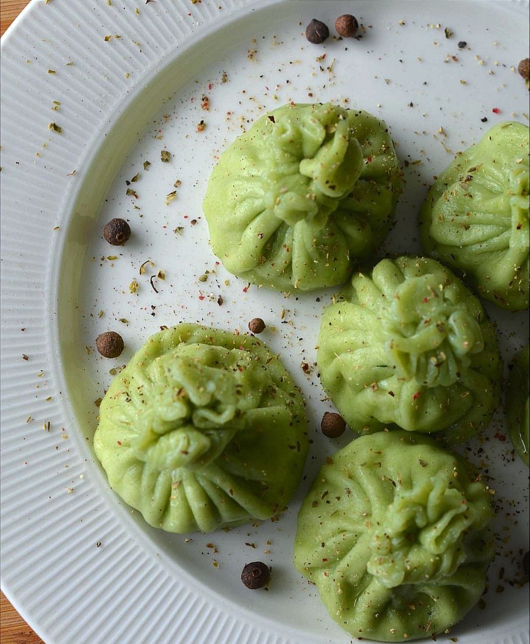 Zielone pierożki w kształcie sakiewek, posypane przyprawami, ułożone na białym, prążkowanym talerzu, z dodatkiem ziarenek pieprzu.